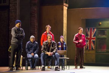 Lavalla on kuuden rennosti pukeutuneen miehen ryhmä, joista osa istuu penkeillä ja osa seisoo. Heidän takanaan on seinä, jossa on Union Jack -lippu ja ovi, mikä viittaa Yhdistyneeseen kuningaskuntaan sijoittuvaan dramaattiseen teatterikohtaukseen.