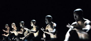 Kuusi paidatonta miestä, joilla on ajeltu pää, istuu rivissä tummaa taustaa vasten, dramaattisen valaistuksen valaisemana, ja suorittavat synkronoituja käsiliikkeitä osana tanssia tai esitystä.