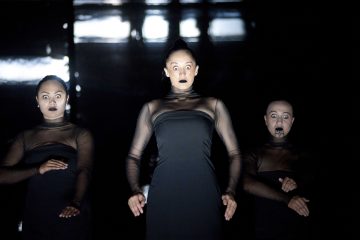 Lavalla esiintyy kolme mustiin läpikuultaviin mekkoihin pukeutunutta naista, joilla on intensiiviset ilmeet, suuret silmät ja dramaattinen musta huulipuna, ja jotka seisovat tummaa taustaa vasten kädet edessään.