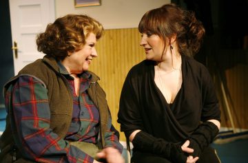 Kaksi naista istuu vierekkäin sisätiloissa hymyillen ja keskustellen keskenään. Toisella on ruudullinen paita ja liivi, ja toinen on pukeutunut mustaan ja hänellä on sormettomat hanskat. Taustalla näkyy suljettu ovi ja puuverhoillut seinät.