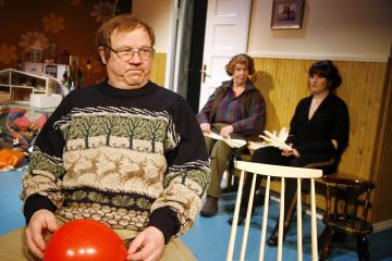 Mies, jolla on silmälasit ja peurakuvioinen villapaita, istuu pitelemässä punaista ilmapalloa, ja hänen takanaan istuu kaksi naista viihtyisässä, lämpimästi valaistussa huoneessa, joka on sisustettu tapetilla ja puupaneelilla.