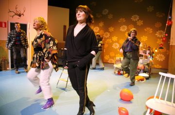 Viisi aikuista tanssii ja hymyilee kirkkaasti valaistussa huoneessa, jossa on keltainen kukkatapetti. Lattialle on ripoteltu punaisia ilmapalloja, ja juhlakoristeet ympäröivät heitä, mikä luo vilkkaan ja juhlavan tunnelman.