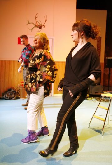 Kolme ihmistä tanssii sisätiloissa sinisellä lattialla. Edessä hymyilee kaksi naista, joista toisella on kukkatakki ja valkoiset housut ja toisella musta asu. Taustalla seisoo mies seinän vieressä, jonka yläpuolelle on asennettu sarvia.