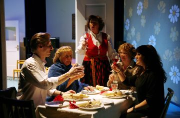 Viisi ihmistä kokoontuu ruokapöydän ympärille, jossa on ruokaa ja viinilaseja. Neljä istuu ja kohottaa lasit maljapuheeseen, kun taas yksi seisoo heidän takanaan kesken puheen tai laulun. Huone on lämpimästi valaistu, ja kukkatapetti on näkyvissä.