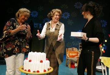 Kolme naista seisoo hymyillen pöydällä olevan valkoisen kuorrutetun kakun ympärillä, jossa on punaisia koristeita. Yksi nainen pitää kädessään pientä valkoista laatikkoa, toinen elehtii vilkkaasti. Taustalla on leikkisä sisustus ja kukkakuvioita.