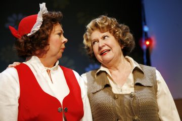 Kaksi pukuun pukeutunutta naista seisoo lähekkäin lavalla. Toisella on valkoinen pusero, punainen liivi ja päähine, toisella beige neuleliivi. He näyttävät keskustelevan vilkkaasti.