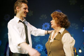 Valkoiseen paitaan ja solmioon pukeutunut nuori mies hymyilee lämpimästi ja ojentaa kätensä kohti vanhempaa kiharatukkaista naista, joka katsoo häntä. Taustalla on kukkakuvioita.