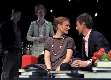 Neljä aikuista hämärässä huoneessa: kaksi istuu lähekkäin punaisella sohvalla ja keskustelee läheisesti, kaksi seisoo heidän takanaan. Etualalla olevalla pöydällä on kirjoja, juomia ja maljakko punaisia tulppaaneja.