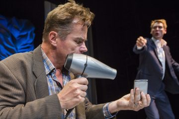 Ruudulliseen paitaan ja bleiseriin pukeutunut mies pitelee hiustenkuivaajaa ja pientä peiliä ja teeskentelee käyttävänsä hiustenkuivaajaa mikrofonina, kun taas toinen pukumies elehtii ja huutaa taustalla lavalla.