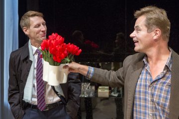 Kaksi miestä seisoo sisätiloissa; toinen pukuun ja takkiin pukeutuneista näyttää huvittuneelta, kun taas toinen, ruudulliseen paitaan ja bleiseriin pukeutunut mies hymyilee ja pitelee valkoista maljakkoa, joka on täynnä kirkkaanpunaisia tulppaaneja, häntä kohti.