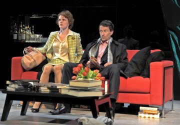 Nainen ja mies istuvat jännittyneinä punaisella sohvalla modernissa olohuoneessa, jota ympäröivät kirjapinot ja sohvapöydällä olevat kukat. Nainen pitelee käsilaukkua ja mies kirjaa, molemmat näyttävät vakavilta.