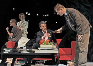 Neljä aikuista on vuorovaikutuksessa dramaattisessa kohtauksessa näyttämöllä; yksi mies kaataa juomaa toiselle punaisella sohvalla, kun kaksi naista istuu taustalla. Heidän edessään on sohvapöydällä kirjoja ja kukkia.