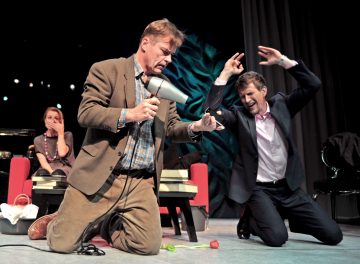 Kaksi pukuihin pukeutunutta miestä esiintyy näyttämöllä dramaattisesti; toinen polvistuu käyttäen hiustenkuivaajaa kengän päällä, kun taas toinen nostaa kätensä ylös hälyttävästi. Taustalla oleva nainen istuu punaisella tuolilla ja peittää suunsa hämmästyneenä.