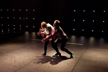 Kaksi tanssijaa esiintyy hämärästi valaistulla näyttämöllä, ja dramaattinen valaistus korostaa heidän ilmeikkäitä asentojaan. Pienet valkoiset valot pistelevät tummaa taustaa, mikä lisää teatterin tunnelmaa.