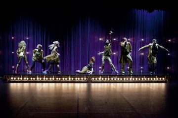 Seitsemän esiintyjää kiiltävissä kultaisissa puvuissa tanssii ja poseeraa tarmokkaasti valaistulla näyttämöllä kimaltelevan violetin verhon edessä, ja yksi henkilö poseeraa lattialla näyttämön keskellä.