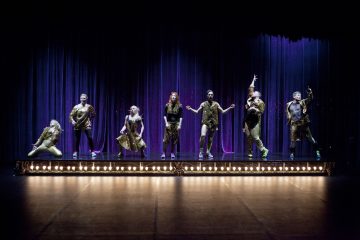 Seitsemän esiintyjää kullanhohtoisissa asuissa poseeraa dramaattisesti valaistulla näyttämöllä, jonka takana on violetit verhot. Kukin henkilö poseeraa ainutlaatuisessa asennossa, ja lavan etuosassa on näyttämövalot.