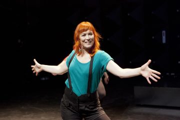 Kirkkaan oranssitukkainen henkilö, jolla on tiilenpunainen paita ja mustat henkselit, hymyilee leveästi kädet ojennettuina tummalla näyttämöllä.