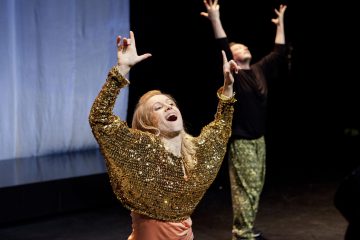 Kullanhohtoiseen toppiin pukeutunut nainen tanssii tarmokkaasti molemmat kädet kohotettuna ja sormet osoittaen. Toinen henkilö, jolla on kuviolliset housut ja musta paita, poseeraa kädet ylhäällä taustalla hämärästi valaistulla lavalla.