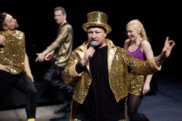 Kultaiseen paljettitakkiin ja silinterihattuun pukeutunut mies laulaa lavalla mikrofoniin, ja hänen takanaan tanssii iloisesti kolme kulta-asuihin pukeutunutta ihmistä ja yksi violetti toppi päässä.