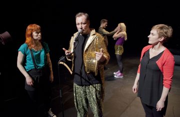 Kultaiseen paljettitakkiin pukeutunut mies puhuu lavalla mikrofoniin, jota reunustaa kolme naista, kun taas kaksi muuta ihmistä on vuorovaikutuksessa taustalla lavan valaistuksen alla.