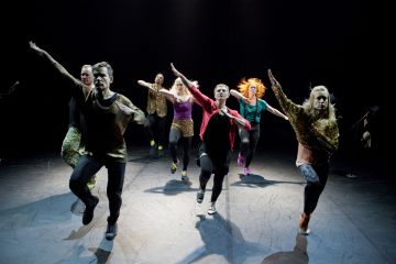 Seitsemän tanssijan ryhmä värikkäissä, rennoissa asuissa esittää synkronoitua tanssirutiinia hämärästi valaistulla näyttämöllä, kädet kohotettuna ja jalat nostettuina energiseen liikkeeseen.