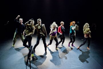 Seitsemän ihmistä tanssii tarmokkaasti hämärästi valaistulla lavalla värikkäisiin ja kiiltäviin vaatteisiin pukeutuneina, ja jokainen heistä tanssii eri poseerauksessa.