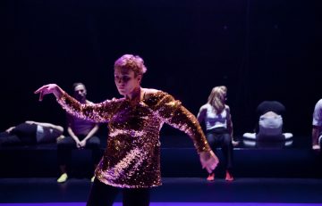 Kultaiseen paljettitoppiin pukeutunut henkilö tanssii dramaattisessa valaistuksessa, kun taas useat muut istuvat tai poseeraavat erilaisissa asennoissa hämärästi valaistussa taustalla.