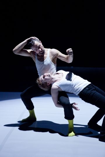 Kaksi tanssijaa, joilla on valkoiset toppahousut, mustat housut ja keltaiset sukat, esiintyvät dramaattisesti; toinen kaartaa taaksepäin, kun toinen seisoo takanaan ja pitelee päätään, molemmat valaisevat pimeän näyttämön valonheittimen valossa.