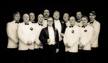 Kaksitoista miestä poseeraa yhdessä muodollisissa puvuissa ja rusettisolmioissa. Yhdellätoista on valkoiset takit ja tummat rusetit, kun taas yhdellä edessä keskellä istuvalla on musta takki ja silmälasit. Tausta on tumma.