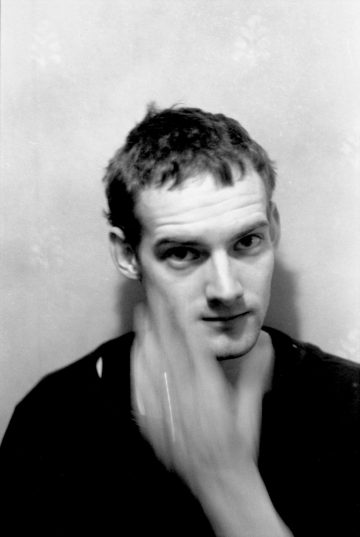 Mustavalkoinen valokuva lyhythiuksisesta miehestä, jolla on tumma paita ja joka katsoo kameraan, hänen kasvonsa ovat osittain hämärtyneet, kun hänen kohotettu kätensä liikkuu hänen edessään. Tausta on tavallinen ja pehmeästi kuvioitu.