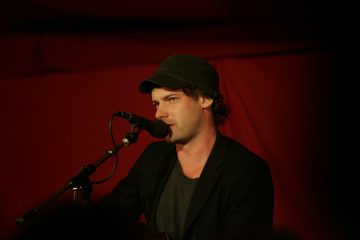 Tummaan takkiin ja lippikseen pukeutunut mies laulaa tai puhuu mikrofoniin lavalla, ja hänen takanaan on punainen tausta.