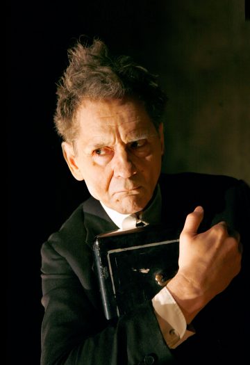 Vanhempi mies, jolla on sotkuinen tukka ja tumma puku, pitää suljettua mustaa kirjaa tiukasti rintaansa vasten ja katsoo sivulle ankaran epäluuloisesti. Tausta on tumma ja dramaattinen valaistus korostaa hänen kasvojaan.