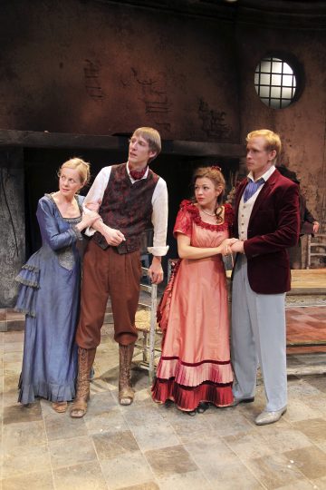 Neljä aikakauden pukuihin pukeutunutta näyttelijää seisoo yhdessä näyttämöllä ja näyttää huolestuneelta. Naisilla on yllään sinisiä ja vaaleanpunaisia röyhelöisiä mekkoja, kun taas miehillä on liivit ja vanhanaikaiset housut. Lavastus on maalaismainen, ja taustalla on pyöreä ikkuna.