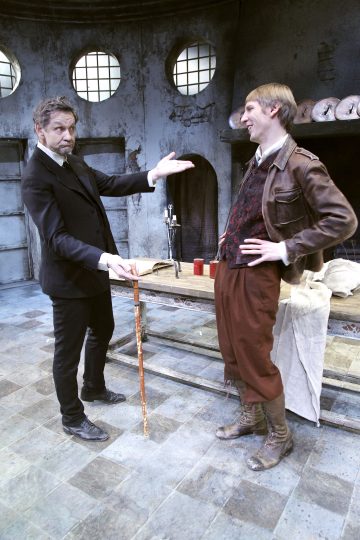 Kaksi näyttelijää näyttämöllä: toinen mustapukuinen näyttelijä viittoo kohti toista, jolla on ruskeat housut, saappaat ja nahkatakki. He seisovat maalaismaisen pöydän ääressä hämärässä teollisuushuoneessa, jossa on pyöreät ikkunat.