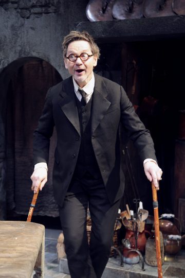 Sisällä seisoo musta pukuinen ja pyöreät silmälasit päässä oleva vanhempi mies, jolla on kaksi puukeppeä kädessään ja yllättynyt ilme kasvoillaan. Taustalla näkyy maalaismainen keittiö, jossa roikkuu kattiloita ja erilaisia astioita.