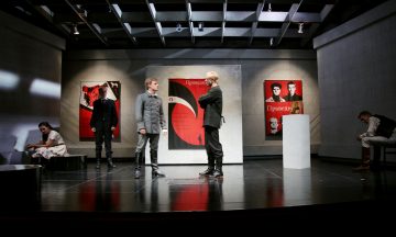 Viisi näyttelijää modernilla näyttämöllä, jonka taustalla on punaisia ja mustia propagandatyylisiä julisteita; kaksi miestä seisoo puhumassa näyttämön keskellä, kun taas kolme muuta istuu sivuilla, mikä luo jännittyneen dramaattisen ilmapiirin.