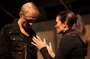 Mies ja nainen seisovat lähekkäin sisätiloissa, molemmat mustiin pukeutuneina. Nainen elehtii käsillään, kun taas mies katsoo alaspäin vaikuttaen vakavalta tai mietteliäältä. Tausta on hämärästi valaistu.