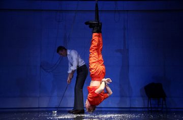 Oranssiin haalariin pukeutunut henkilö roikkuu ylösalaisin kädet selän taakse sidottuina, kun toinen valkoiseen paitaan ja solmioon pukeutunut henkilö pelaa golfia vieressä hämärästi valaistulla lavalla.