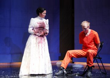 Veren tahrimassa valkoisessa mekossa oleva nainen seisoo oranssiin vankilapukuun pukeutuneen miehen vieressä, joka istuu tuolilla kädet selän taakse sidottuina. Kohtaus sijoittuu hämärästi valaistulle näyttämölle.