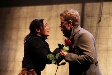 Nainen ja mies keskustelevat intensiivisesti näyttämöllä; nainen pitää kädessään punaista ruusua ja näyttää ahdistuneelta, kun taas harmaaseen takkiin pukeutunut mies katsoo häntä tarkasti dramaattisessa valaistuksessa.