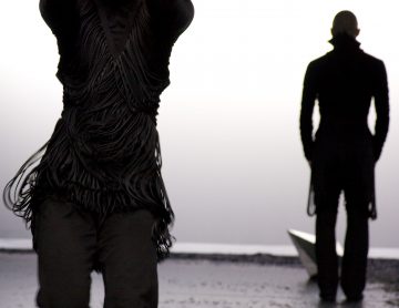 Kaksi tummiin vaatteisiin pukeutunutta ihmistä seisoo rannalla; toinen on etualalla kädet kohotettuna, ja toinen seisoo kauempana veden äärellä lähellä paperivenettä. Kohtaus on tunnelmallinen ja sumuinen.