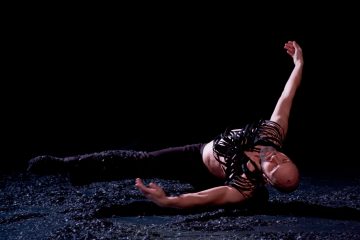 Kalju tanssija mustassa, olkaimellisessa puvussa makaa kyljellään tummalla, kuvioidulla näyttämöllä, toinen käsi ojennettuna pään yläpuolelle ja toinen ojennettuna, luoden dramaattisen ja ilmeikkään poseerauksen.