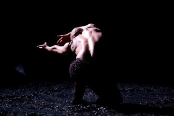Henkilö, jolla on paljas selkä, polvistuu pimeällä näyttämöllä, kädet dramaattisesti ojennettuina ja pää kumartuneena, ja häntä valaisee yksi valonlähde mustaa taustaa vasten.