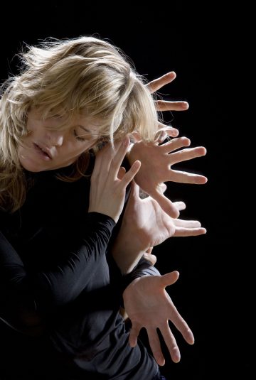 Vaaleatukkainen nainen mustassa asussa poseeraa mustaa taustaa vasten. Useat kädet ympäröivät hänen päätään ja hartioitaan, mikä luo kerroksellisen, surrealistisen vaikutelman, ikään kuin hänellä olisi monta kättä.