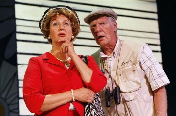 Punaiseen mekkoon ja helmiin pukeutunut nainen seisoo mietteliäänä lippalakkipäisen miehen vieressä, jolla on kiikarit kaulassaan. Molemmat katsovat keskittyneesti eteenpäin, ikään kuin reagoivat johonkin näyttämön ulkopuolella tapahtuvaan, mikä viittaa dramaattiseen tai komedialliseen kohtaukseen.