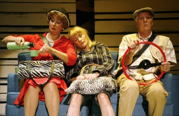 Kolme ihmistä istuu sinisellä penkillä, poseerattuna kuin autossa. Vasemmalla istuva nainen kaataa juomaa, keskellä istuva nainen nukkuu olkapäällään, ja oikealla istuva mies teeskentelee ohjaavansa autoa punaisella leluohjauspyörällä.