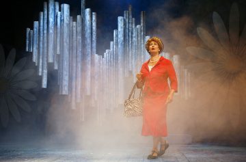 Punapukuinen ja helmiin pukeutunut henkilö seisoo lavalla sumussa, kädessään seepra-raidallinen laukku. Taustalla on suuria päivänkakkaroita ja pystysuoria putkikoristeita, jotka luovat dramaattisen teatterikohtauksen.