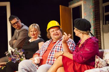 Neljä aikuista istuu kukkasohvalla keskustellen. Yhdellä miehellä on harmaa puku ja silmälasit, toisella ruudulliseen paitaan pukeutuneella miehellä on keltainen rakennuskypärä. Kahdella naisella on mekko, toisella vaaleanpunainen ja musta baretti.