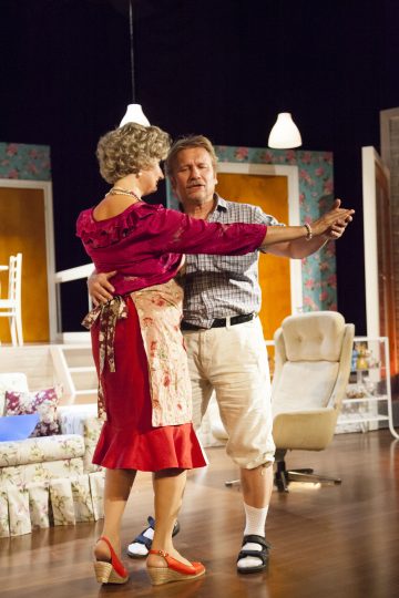 Lavalla tanssii kaksi näyttelijää, joista toisella on punainen mekko, harmaa peruukki ja esiliina, kun taas toisella on ruudullinen paita, shortsit ja sandaalit. Viihtyisässä lavastuksessa on kukkakalusteita ja lämmin valaistus.