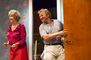 Hymyilevä vanhempi nainen punaisessa mekossa ja mies ruudullisessa paidassa ja khakishortseissa seisovat kukkatapetoidussa huoneessa; mies pitää puista ovea auki; molemmat vaikuttavat iloisilta ja keskustelevilta.